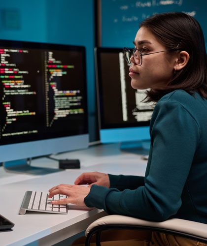 Mujer con gafas escribiendo en un teclado frente a dos pantallas con líneas de código